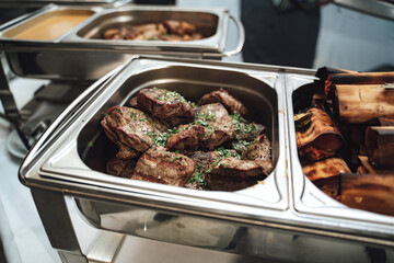 Row of stainless hotel pans on food warmers with various meals. Juicy beef steaks. Self-service buffet table. Celebration, party, birthday or wedding