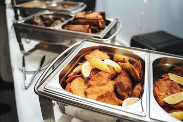 Row of stainless hotel pans on food warmers with various meals. Fried schnitzel with slices of...