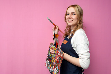 Young happy girl artist holds drawing equipment and draws with a brush on a pink background, the...