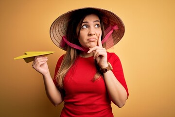 Tourist woman wearing traditional asian rice paddy straw hat holding paper plane for a trip serious face thinking about question, very confused idea