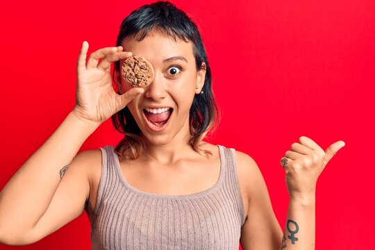 Young woman holding cookie pointing thumb up to the side smiling happy with open mouth