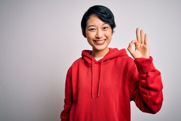 Young beautiful asian girl wearing casual sweatshirt with hoodie over white background smiling positive doing ok sign with hand and fingers. Successful expression.