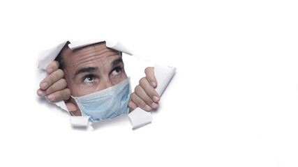 mid age caucasian man wearing protective mask or surgeon breaking through a white paper sheet or wall watching outside in confinement for coronavirus covid-19