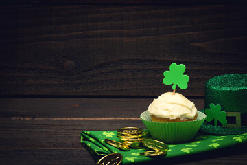 Happy St. Patricks Day Still Life with a Cupcake, Shamrocks, Hat, and Gold Coins for Good Luck and Celebration.  It's a horizontal photo with dark wood background