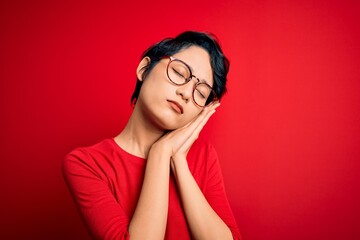 Young beautiful asian girl wearing casual t-shirt and glasses over isolated red background sleeping tired dreaming and posing with hands together while smiling with closed eyes.