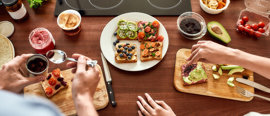 Top view of couple of vegetarians preparing various healthy toasts in the kitchen together. Man getting out sun dried tomatoes while woman adding chia seeds
