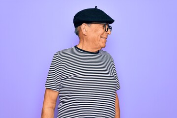 Senior handsome hoary man wearing french beret and glasses standing over purple background looking away to side with smile on face, natural expression. Laughing confident.