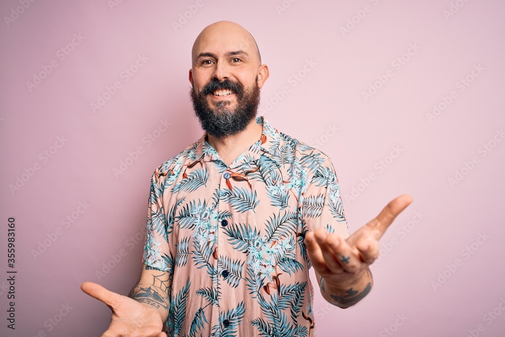 Wall mural Handsome bald man with beard and tattoo wearing casual floral shirt over pink background smiling cheerful with open arms as friendly welcome, positive and confident greetings