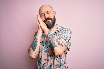 Handsome bald man with beard and tattoo wearing casual floral shirt over pink background sleeping tired dreaming and posing with hands together while smiling with closed eyes.