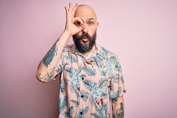 Handsome bald man with beard and tattoo wearing casual floral shirt over pink background doing ok gesture shocked with surprised face, eye looking through fingers. Unbelieving expression.