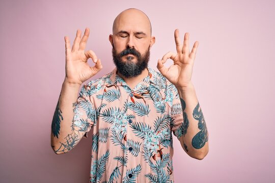 Handsome bald man with beard and tattoo wearing casual floral shirt over pink background relaxed and smiling with eyes closed doing meditation gesture with fingers. Yoga concept.