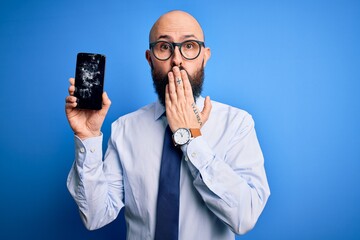 Handsome bald business man with beard holding broken smartphone showing cracked screen cover mouth with hand shocked with shame for mistake, expression of fear, scared in silence, secret concept