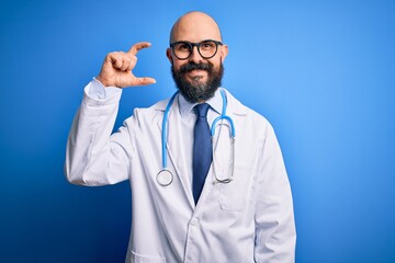 Handsome bald doctor man with beard wearing glasses and stethoscope over blue background smiling and confident gesturing with hand doing small size sign with fingers looking and the camera. Measure