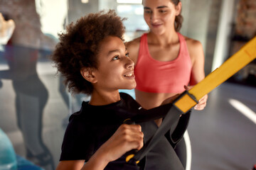 Portrait of teenage boy training using fitness straps in gym while female trainer helping him....