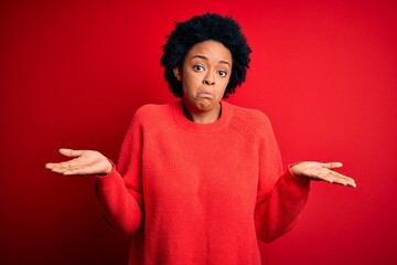 Young beautiful African American afro woman with curly hair wearing casual sweater clueless and confused expression with arms and hands raised. Doubt concept.