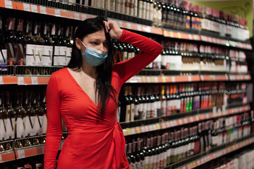 Beautiful  brunette girl   stands near the shelves with bottle of alcohol in the supermarket. She is dressed in a  red dress and medical protective  mask