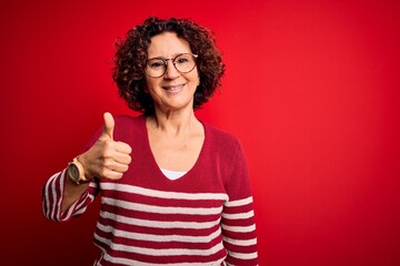 Middle age beautiful curly hair woman wearing casual striped sweater over red background doing happy thumbs up gesture with hand. Approving expression looking at the camera showing success.