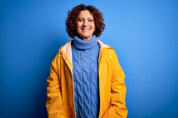 Middle age curly hair woman wearing rain coat standing over isolated blue background with a happy and cool smile on face. Lucky person.