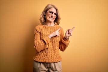 Middle age beautiful blonde woman wearing casual sweater and glasses over yellow background Pointing aside worried and nervous with both hands, concerned and surprised expression