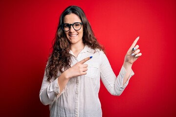 Young beautiful woman with curly hair wearing shirt and glasses over red background smiling and looking at the camera pointing with two hands and fingers to the side.