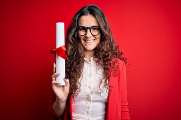 Young beautiful woman with curly hair holding university diploma degree over red background with a happy face standing and smiling with a confident smile showing teeth - obrazy, fototapety, plakaty