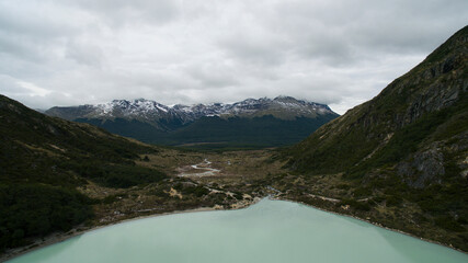 Aerial view of glacier water Emerald Lake in the mountaintop in Ushuaia, Tierra del Fuego, Patagonia Argentina. A stream flows downhill the forest and valley into the Andes mountains with snowy peaks