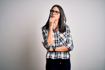 Young brunette woman with blue eyes wearing casual shirt and glasses over white background with hand on chin thinking about question, pensive expression. Smiling with thoughtful face. Doubt concept.