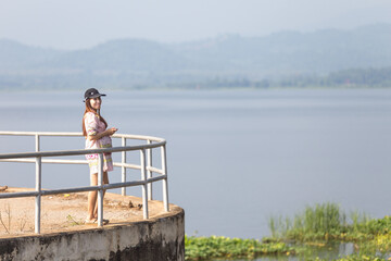 young pregnant woman standing on a pier at the lake in mountain side during beautiful sunny day in summer season