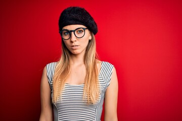 Beautiful blonde woman with blue eyes wearing french beret and glasses over red background with serious expression on face. Simple and natural looking at the camera.