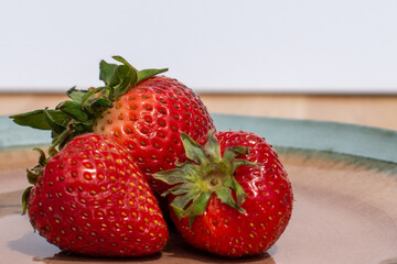 Strawberries on a decorative glass plate.  The plate is sitting on a wooden oak table with a green background. There are three fresh strawberries of various sizes, bright red color, and juicy texture.