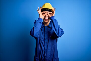 Young beautiful worker woman with blue eyes wearing security helmet and uniform doing ok gesture like binoculars sticking tongue out, eyes looking through fingers. Crazy expression.