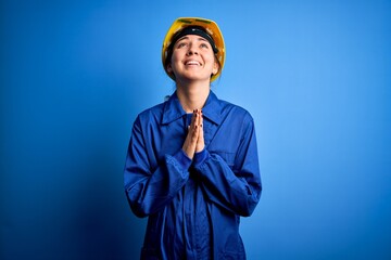 Young beautiful worker woman with blue eyes wearing security helmet and uniform begging and praying with hands together with hope expression on face very emotional and worried. Begging.