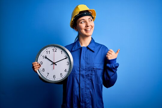 Beautiful blonde worker woman with blue eyes wearing security helmet holding big clock pointing and showing with thumb up to the side with happy face smiling