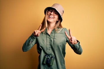 Beautiful blonde explorer woman with blue eyes wearing hat and glasses using binoculars smiling looking to the camera showing fingers doing victory sign. Number two.