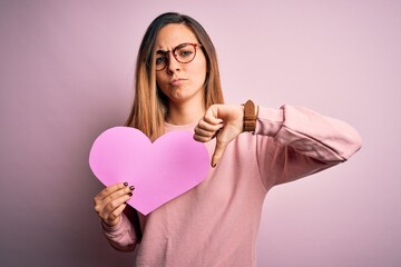 Young beautiful blonde romantic woman wearing glasses holding big pink heart with angry face, negative sign showing dislike with thumbs down, rejection concept