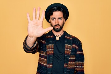 Naklejka premium Young handsome hispanic bohemian man wearing hippie style and boho hat doing stop sing with palm of the hand. Warning expression with negative and serious gesture on the face.
