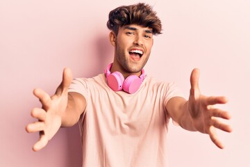 Young hispanic man listening to music using headphones looking at the camera smiling with open arms for hug. cheerful expression embracing happiness.