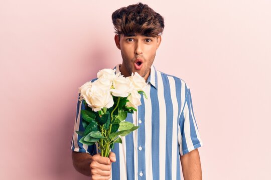 Young hispanic man holding flowers scared and amazed with open mouth for surprise, disbelief face