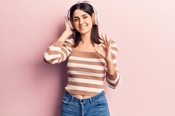 Young beautiful girl listening to music using headphones doing ok sign with fingers, smiling friendly gesturing excellent symbol