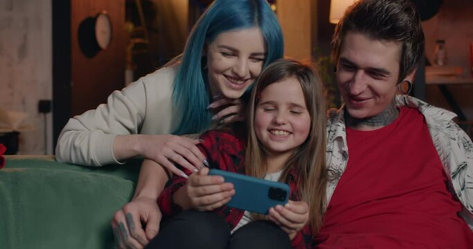 Young family laughing and touching screen while using smartphone at home. Modern mom, dad and their pretty daughter having good time together while sitting on sofa . Concept of leisure.