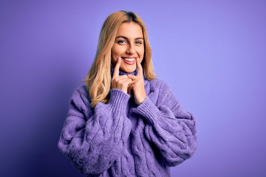 Young beautiful blonde woman wearing casual turtleneck sweater over purple background Smiling with open mouth, fingers pointing and forcing cheerful smile