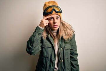 Young brunette skier woman wearing snow clothes and ski goggles over white background pointing unhappy to pimple on forehead, ugly infection of blackhead. Acne and skin problem