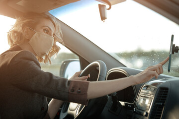 Fototapeta premium A beautiful girl driver with a mask on her face, sitting behind the wheel of a car uses the Navigator in the daytime.