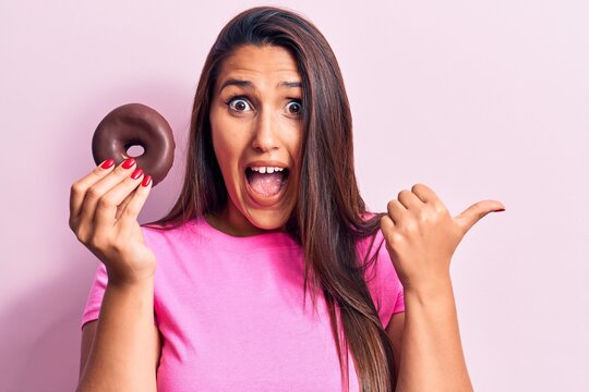 Young beautiful brunette woman holding chocolate donut pointing thumb up to the side smiling happy with open mouth