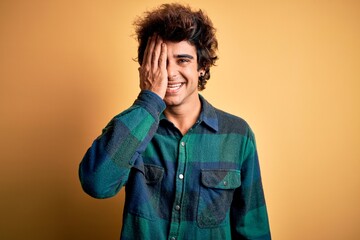 Young handsome man wearing casual shirt standing over isolated yellow background covering one eye with hand, confident smile on face and surprise emotion.