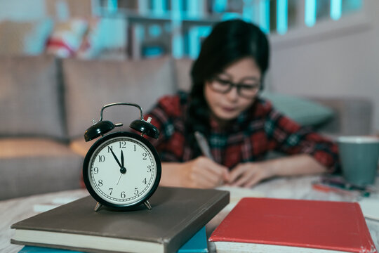 Closeup Of A Clock Against Blurred Background Of A Diligent Japanese Girl Burning Midnight Oil. Asian Female Student Still Up Studying Late At Night