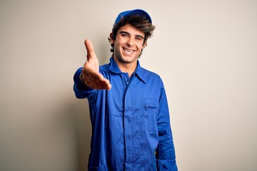 Young mechanic man wearing blue cap and uniform standing over isolated white background smiling friendly offering handshake as greeting and welcoming. Successful business.
