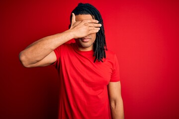 Young handsome african american afro man with dreadlocks wearing red casual t-shirt covering eyes with hand, looking serious and sad. Sightless, hiding and rejection concept