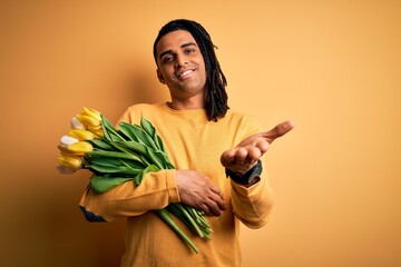 Young african american afro romantic man with dreadlocks holding bouquet of yellow tulips smiling cheerful offering palm hand giving assistance and acceptance.