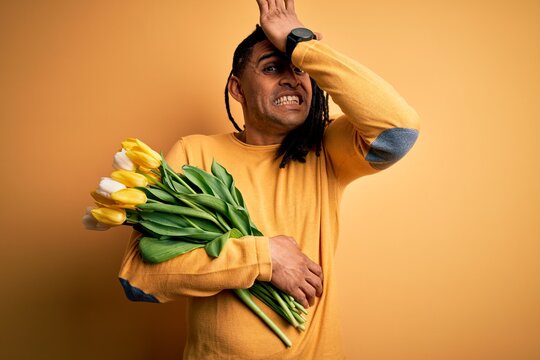 Young african american afro romantic man with dreadlocks holding bouquet of yellow tulips surprised with hand on head for mistake, remember error. Forgot, bad memory concept.